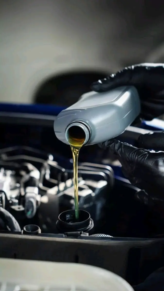 Mãos com luvas de proteção despejam óleo lubrificante automotivo a partir de uma embalagem plástica diretamente no bocal do motor do veículo.